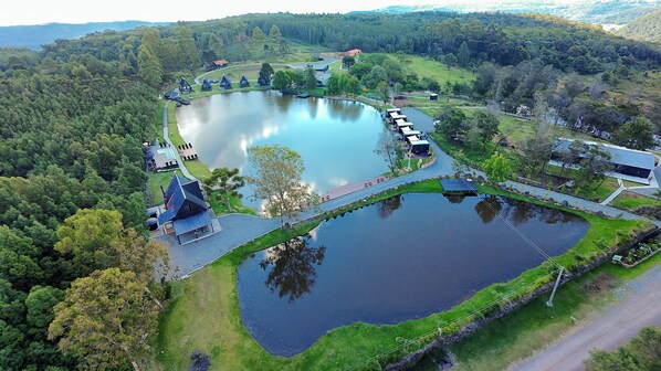 An aerial view of a lakeside property with a large pond, a bridge, and several buildings including a house with a steep roof and a smaller structure with a sloped roof. - Vila Gramado (Gramado)