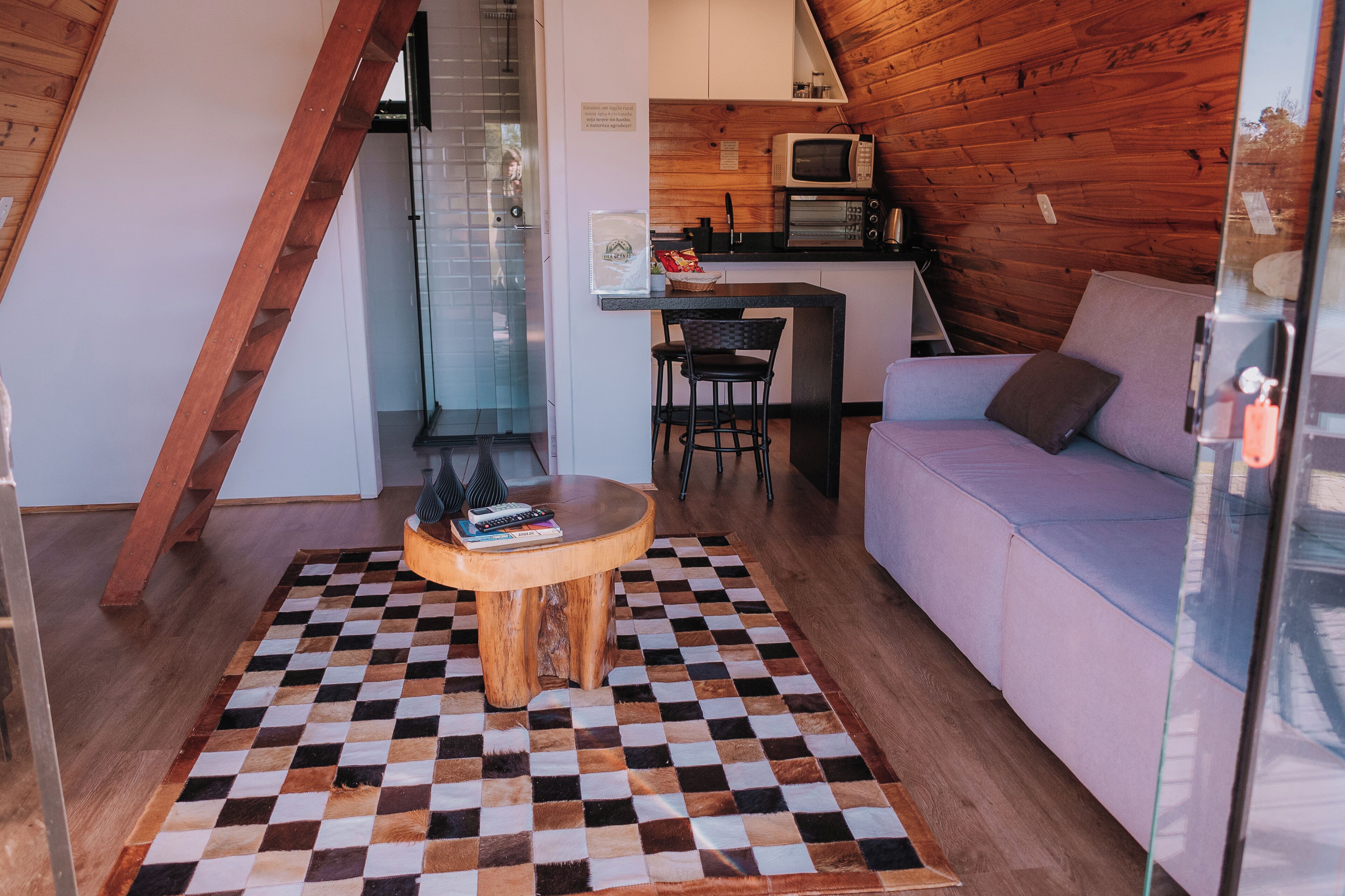 Un espacio habitable compacto con piso a cuadros, un sofá, una mesita con libros, una zona de cocina con microondas y una escalera.