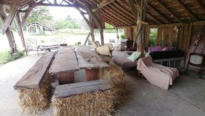 A rustic wooden structure with a large table and hay bales, surrounded by seating and a view of greenery. - FARMHOUSE GITE WITH SHARED POOL - A CHANGE OF SCENERY FOR FAMILY AND FRIENDS (Cachen)