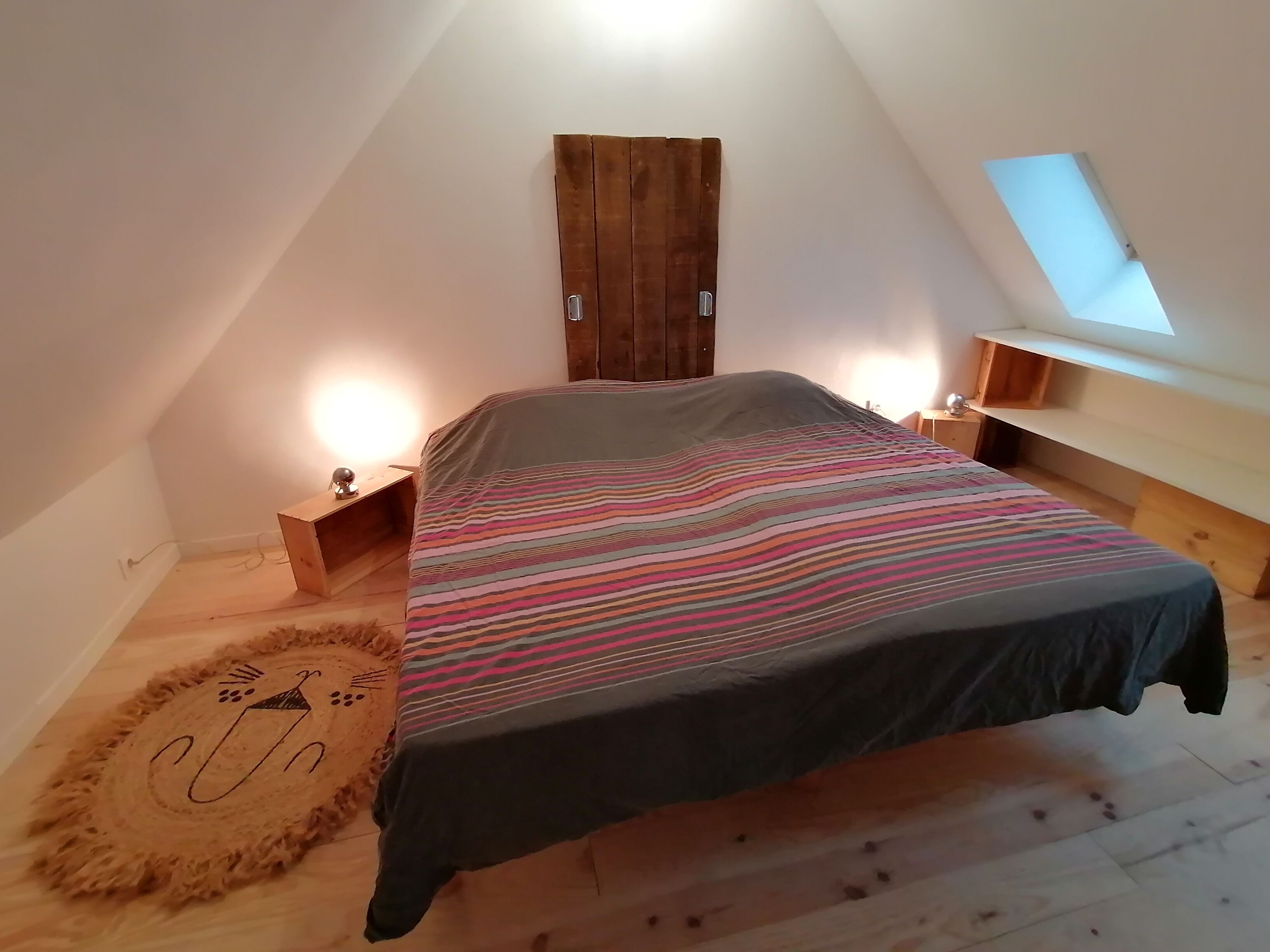 Un dormitorio con techo inclinado, cama con colcha a rayas, armario de madera, escritorio pequeño con lámpara y tragaluz.