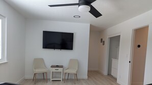 Une chambre à coucher avec un lit, un téléviseur fixé au mur, deux chaises, une petite table et un ventilateur de plafond.