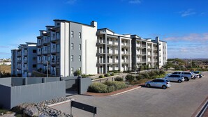 A multi-story residential building with balconies, a parking area with several cars, and a clear blue sky. - NIVICA Lifestyle Living 20, Langebaan 4-Sleeper (Langebaan)