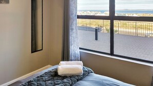 A hotel room with a large window, a bed with a grey blanket, and a white towel on the bed. - NIVICA Lifestyle Living 20, Langebaan 4-Sleeper (Langebaan)