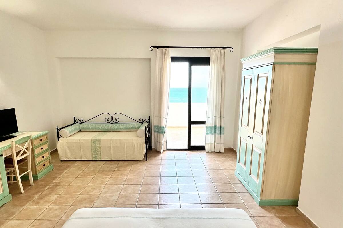 Ein Schlafzimmer mit Bett, Schreibtisch, Kleiderschrank und Meerblick durch eine Glastür.