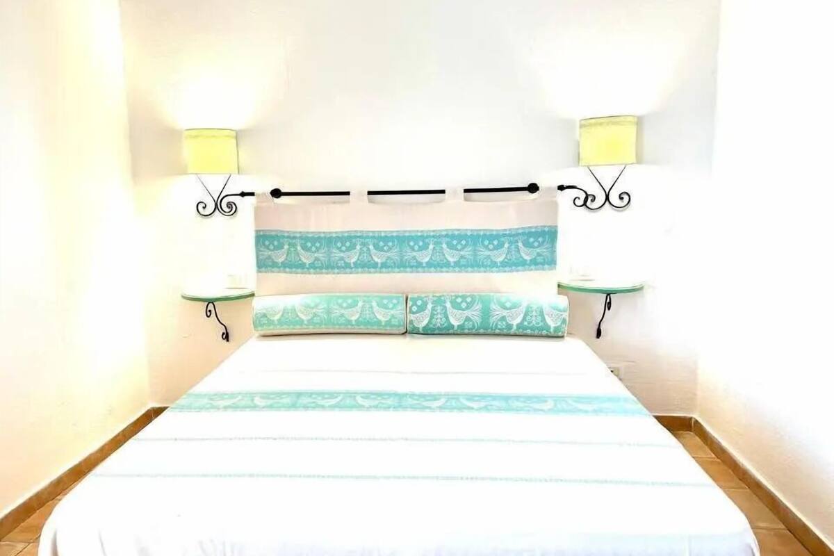 Ein Bett mit weißer und türkiser Bettdecke, zwei passende Kissen und ein Kopfteil mit dekorativem Muster. Auf beiden Seiten des Bettes stehen jeweils Nachttische mit Lampen.