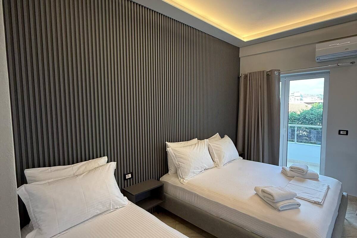 A modern hotel room with two single beds, a large window with curtains, and a ceiling light fixture.