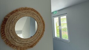 A room with a round woven mirror on the wall, a window with a view of greenery, and a door with an exit sign. - Shehu Villa Ksamil (Ksamil)