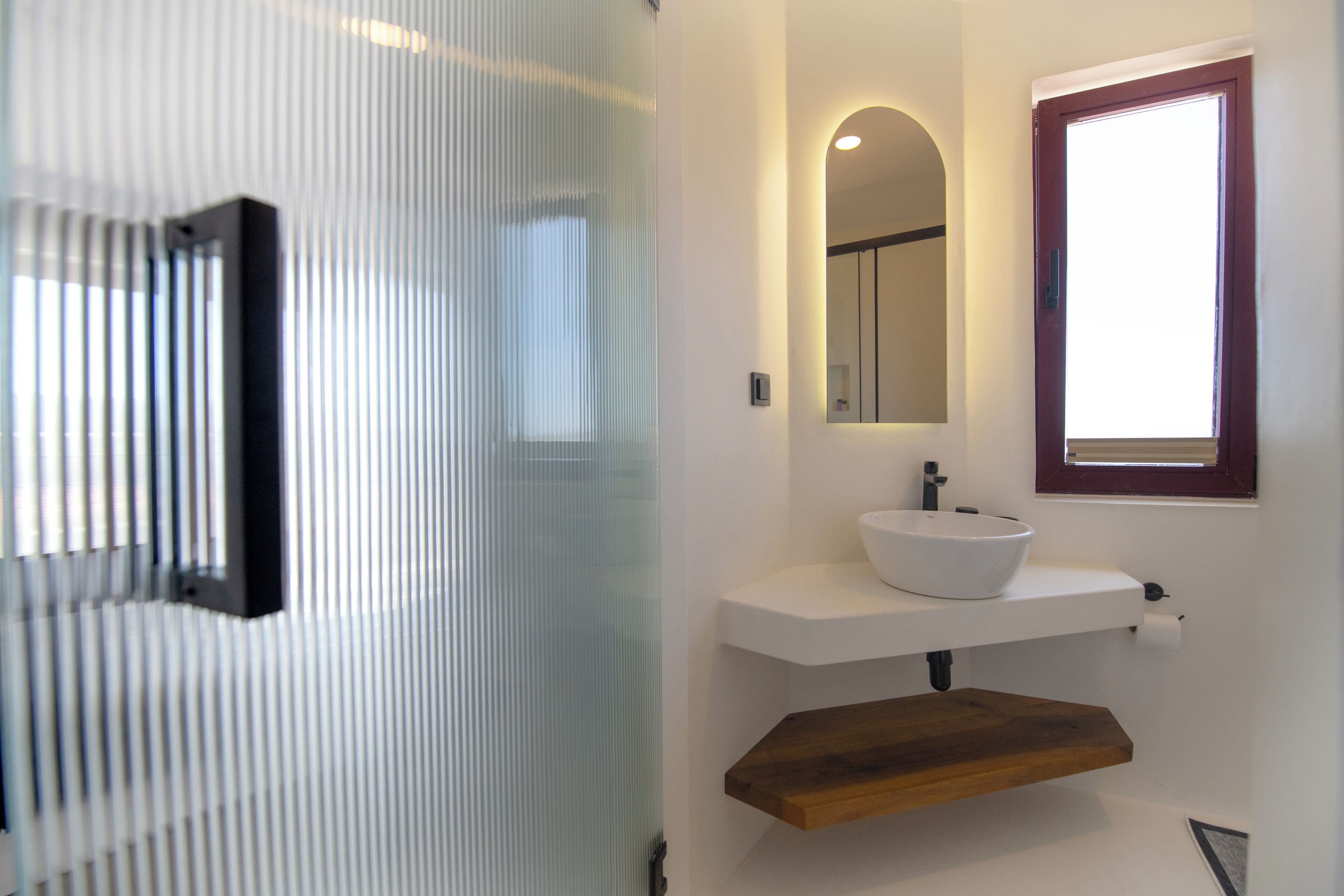 A modern bathroom with a white sink, a wooden shelf, a mirror, and a frosted glass door.
