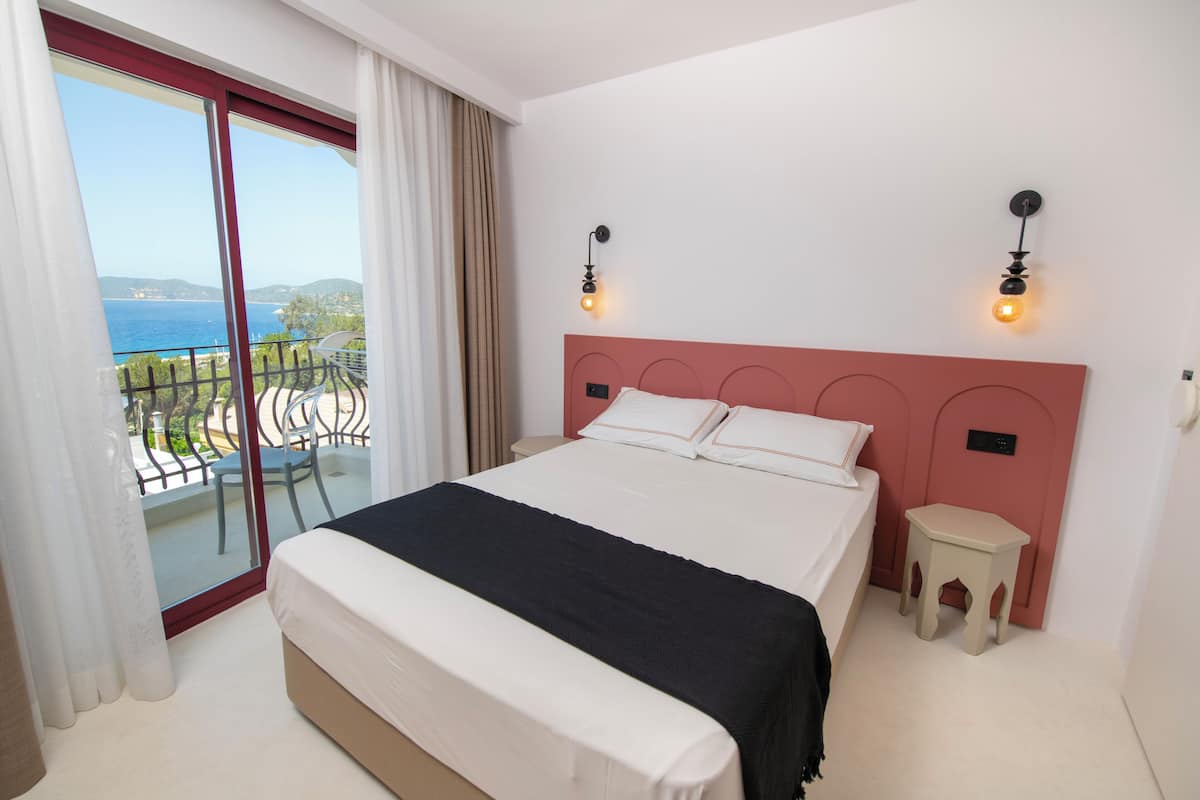 Un dormitorio con una cama grande, un taburete pequeño y vista al mar desde un balcón.