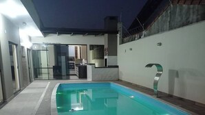 A rectangular swimming pool with a tiled deck, surrounded by a modern building with glass doors and a kitchen area visible through the glass. - House with HEATED POOL (No additional cost) (Bopiranga Itanhaém)