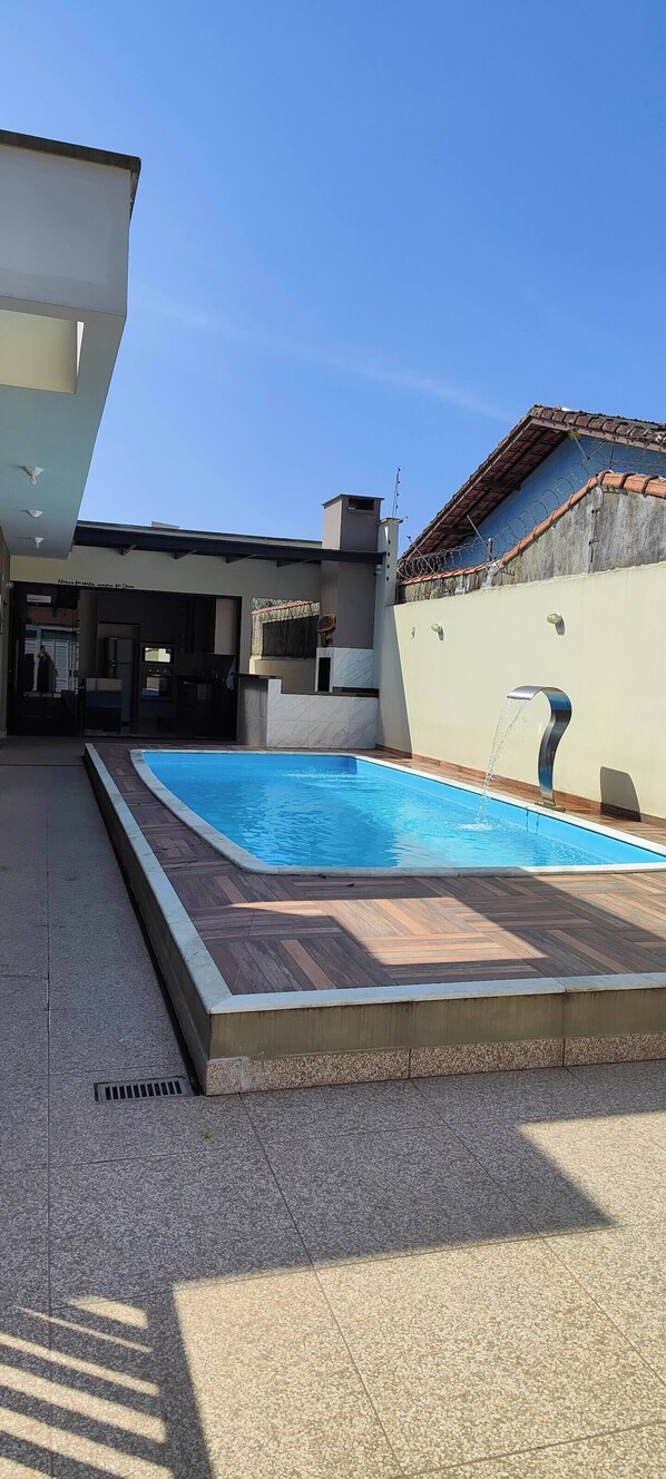 Una piscina con bordo piastrellato, una zona coperta con tetto e un edificio con ingresso visibile.