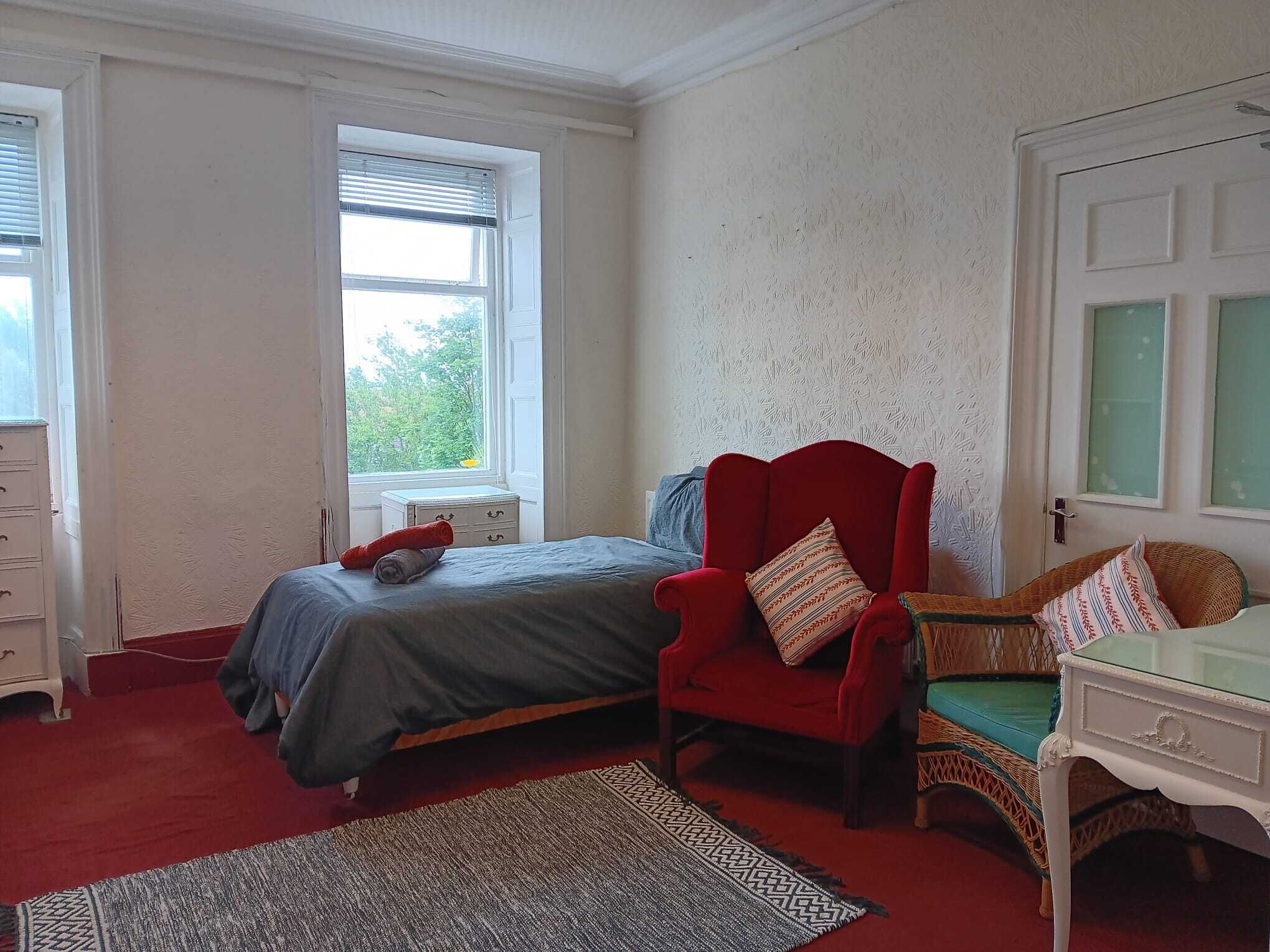 Une chambre à coucher avec un lit, un fauteuil rouge, une table d’appoint en osier et une fenêtre avec des stores.