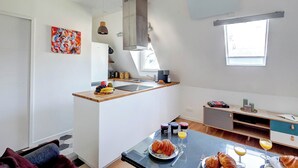 A modern kitchen with a central island, a dining area with a table set for breakfast, and a large window providing natural light. - Champs-Élysées - 1BR (Paris)
