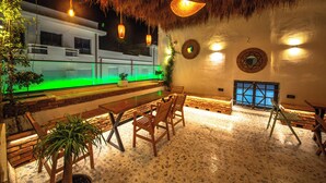 An outdoor dining area with a thatched roof, wooden furniture, and a view of a building across a water feature. - Vega 43 Hostel (Sarande)
