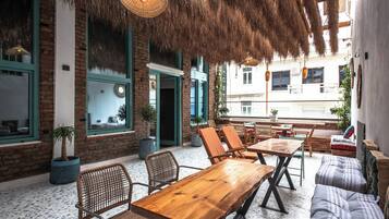 An outdoor seating area with wooden tables and chairs, a thatched roof, and a view of the street.