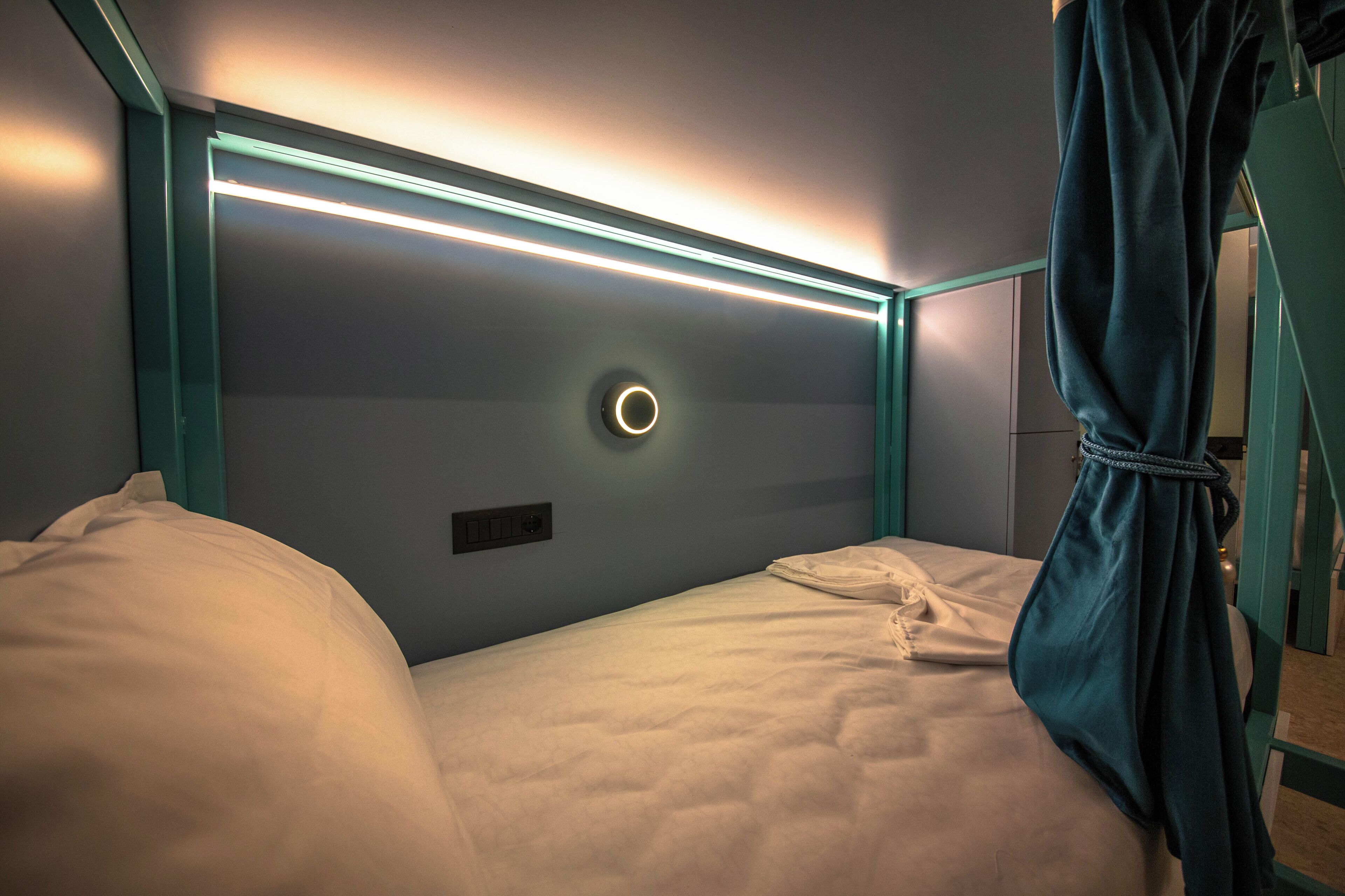 A single bed with a headboard featuring a circular light fixture and a bedside table with a power outlet.