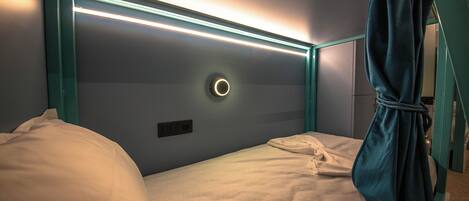 A single bed with a headboard featuring a circular light fixture and a bedside table with a power outlet.