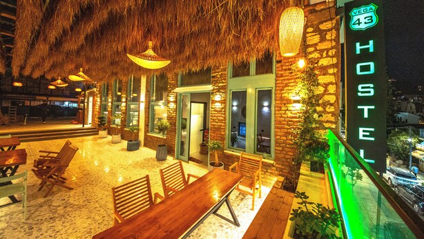 A cozy outdoor seating area with wooden tables and chairs, a thatched roof, and a neon sign indicating a hostel. - Vega 43 Hostel (Sarande)