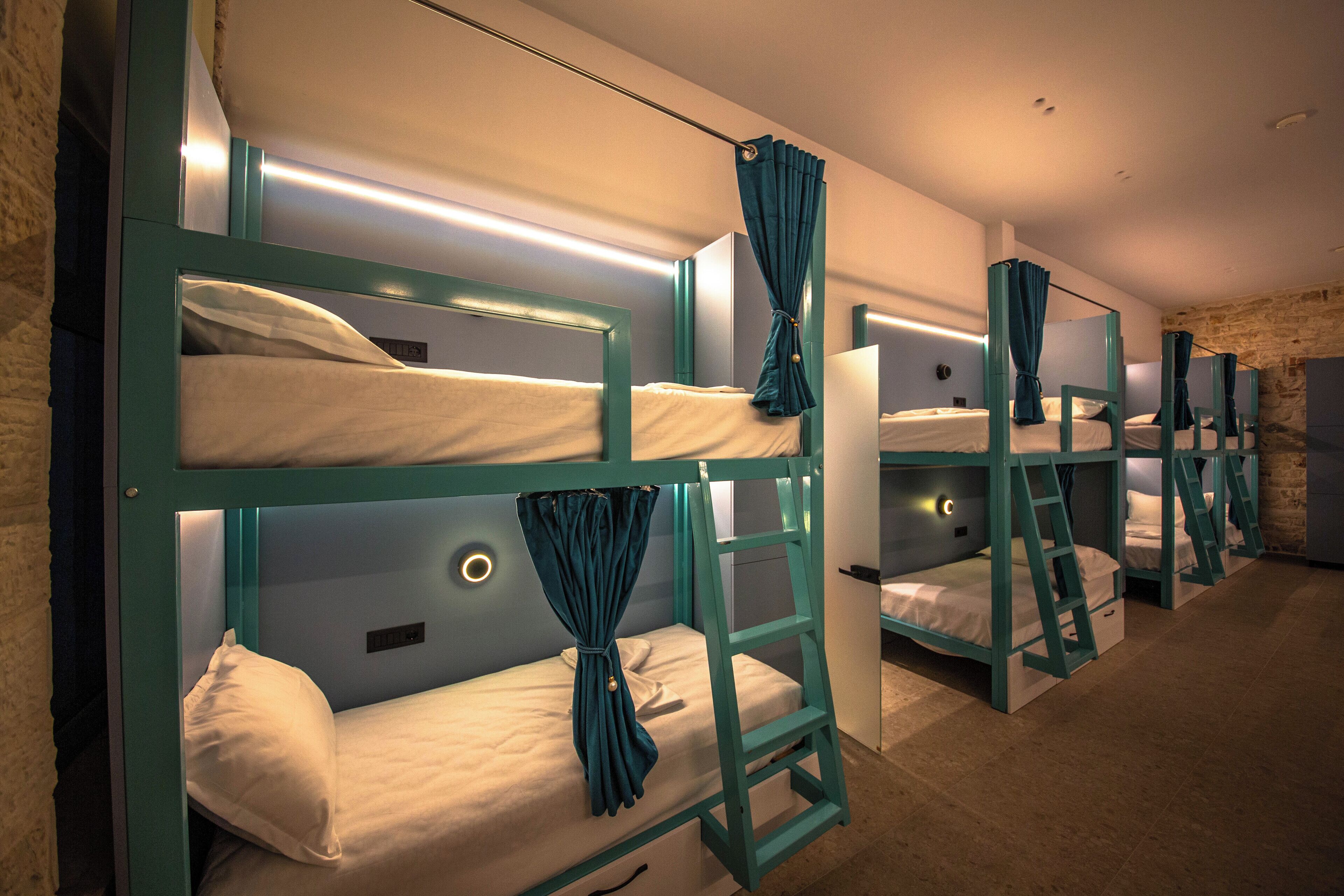 A row of bunk beds with teal frames and white bedding, arranged in a corridor with stone walls and a warm light source.