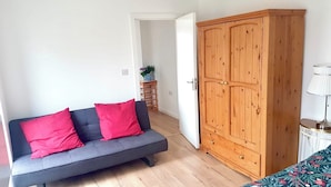 Una habitación con un sofá gris, cojines rojos, un armario de madera, una mesita con una lámpara y una puerta que da a otra habitación.