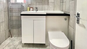 Un baño moderno con inodoro blanco, lavamanos y espejo sobre él. Hay un perchero y un cepillo de inodoro en el suelo.
