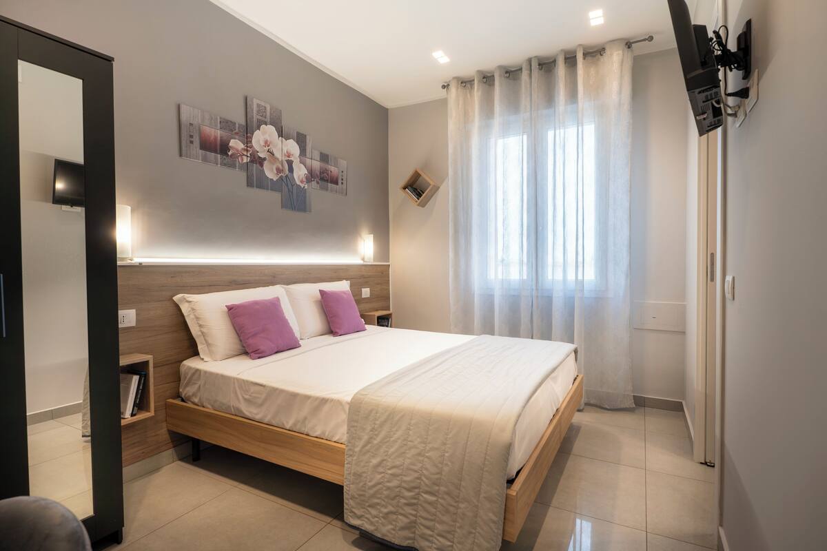 A modern bedroom with a wooden bed, a bedside table, a wall-mounted TV, and a window with sheer curtains.