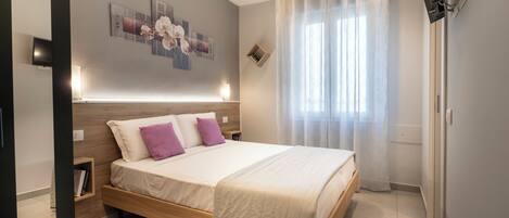 A modern bedroom with a wooden bed, a bedside table, a wall-mounted TV, and a window with sheer curtains.