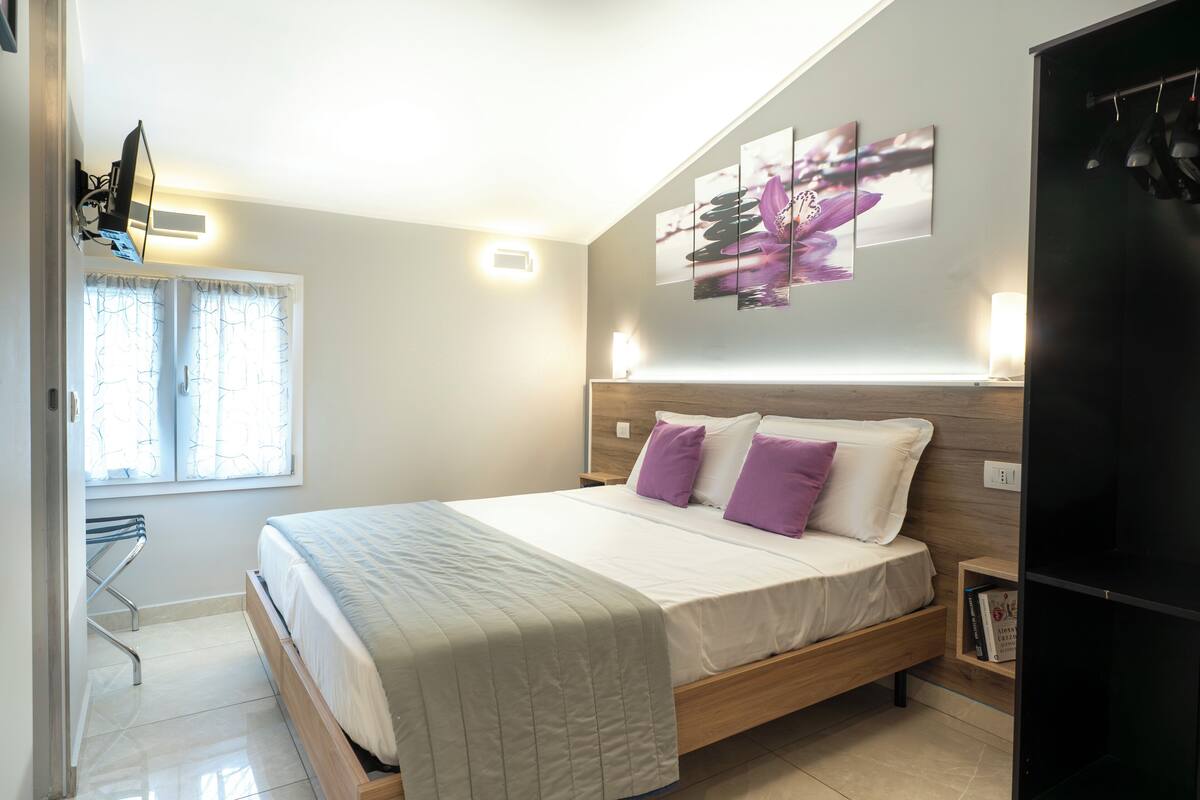 A bedroom with a bed, a nightstand, a wall-mounted TV, and a window with curtains.