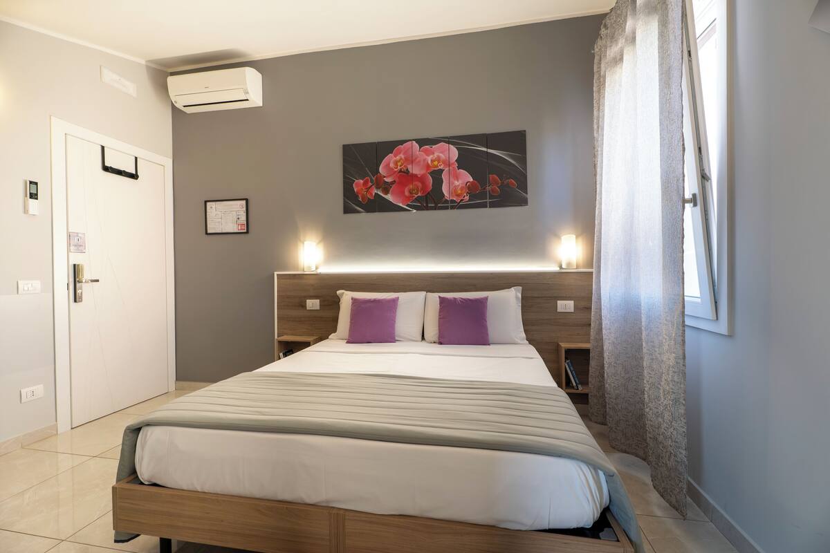 A neatly made bed with a wooden frame and white bedding, a wall-mounted air conditioner, a framed artwork of pink flowers, and a window with curtains.