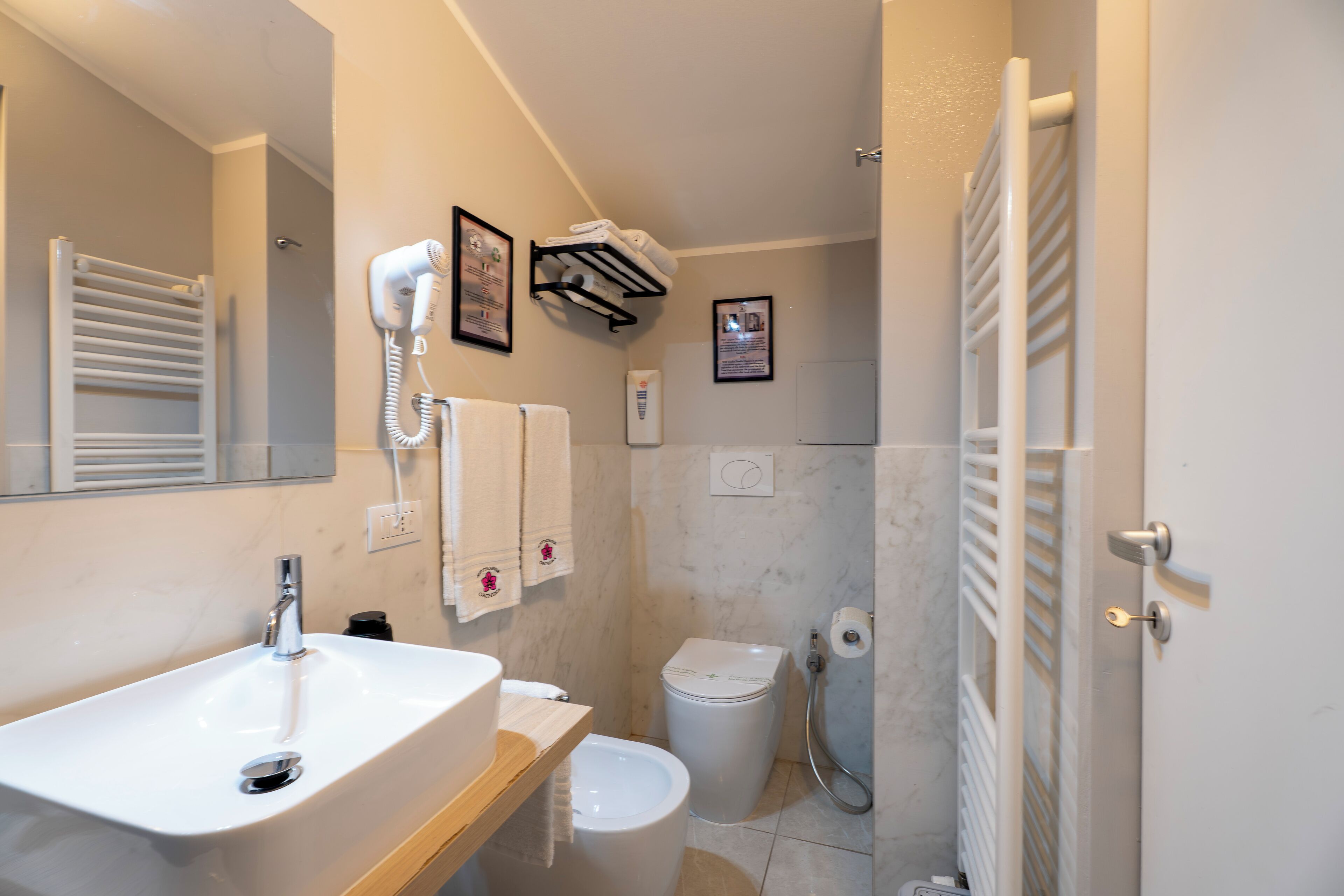 Ein Badezimmer mit Waschbecken, Toilette und Dusche. Handtücher und ein Fön sind an der Wand angebracht.