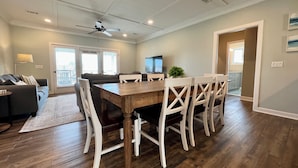 Un área de comedor con una mesa de madera y sillas blancas, un ventilador de techo y un televisor al fondo.