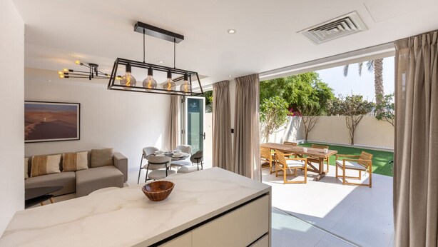 A modern kitchen with a marble countertop, a dining area with wooden furniture, and a view of a garden through large sliding doors. - Arabian Ranches 1 - Upgraded Three Bedroom Villa (Arabian Ranches)