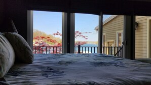 Un dormitorio con un ventanal grande que da a un balcón con vistas a un cuerpo de agua y follaje otoñal.
