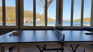 Mesa de ping pong con dos paletas, un cesto y una raqueta de tenis de mesa, ubicada cerca de amplias ventanas con vistas al agua y árboles.