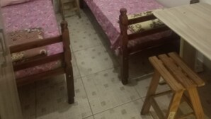 A small, simple room with two wooden beds, a small table, and a standing fan.