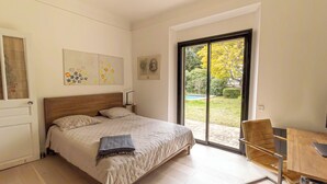 Een moderne slaapkamer met een groot bed, een houten stoel en uitzicht op een tuin via een glazen schuifdeur.