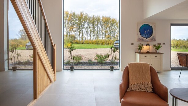 A spacious room with a large window offering a view of a garden and trees. There is a wooden staircase on the left, a brown leather chair in the center, and a white cabinet with a lamp on the right. - Beautiful Barn Conversion - Orchard Escape for 12 (Cropthorne)