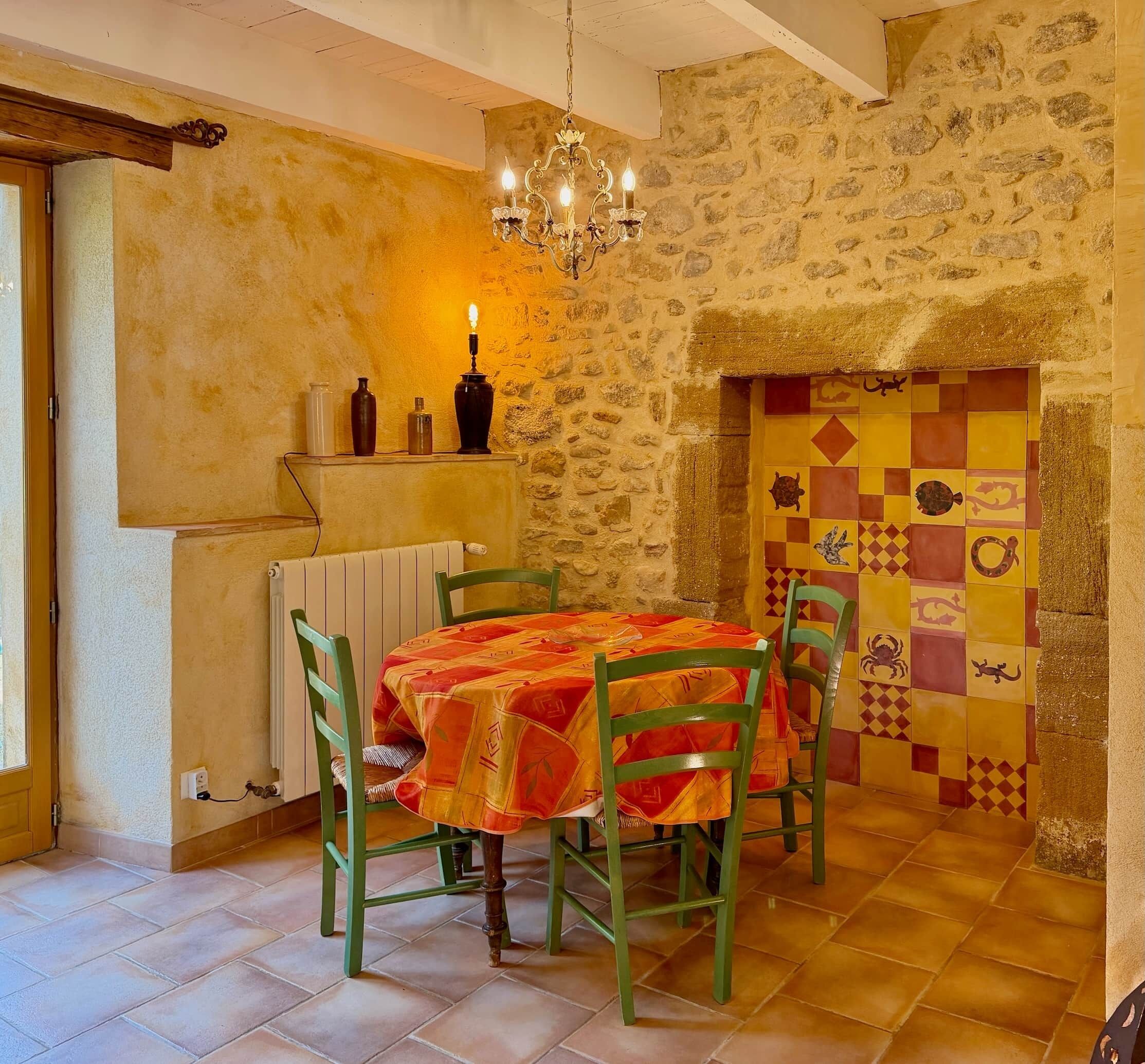 A dining area with a table covered in a patterned tablecloth, green chairs, a chandelier, and a stone wall with a decorative tile design.