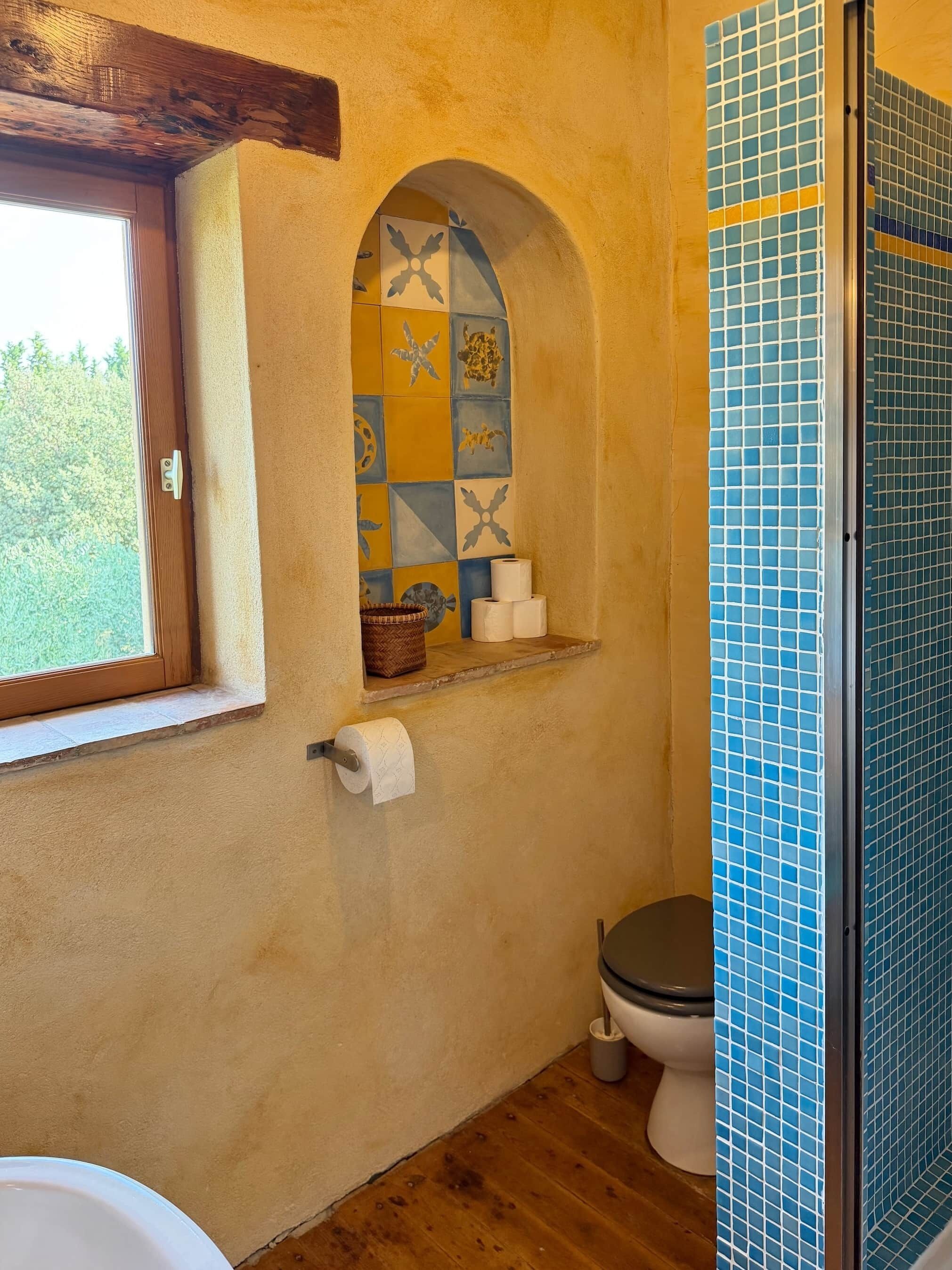 A bathroom with a wooden floor, a window, a tiled shower enclosure, and a shelf with toiletries.