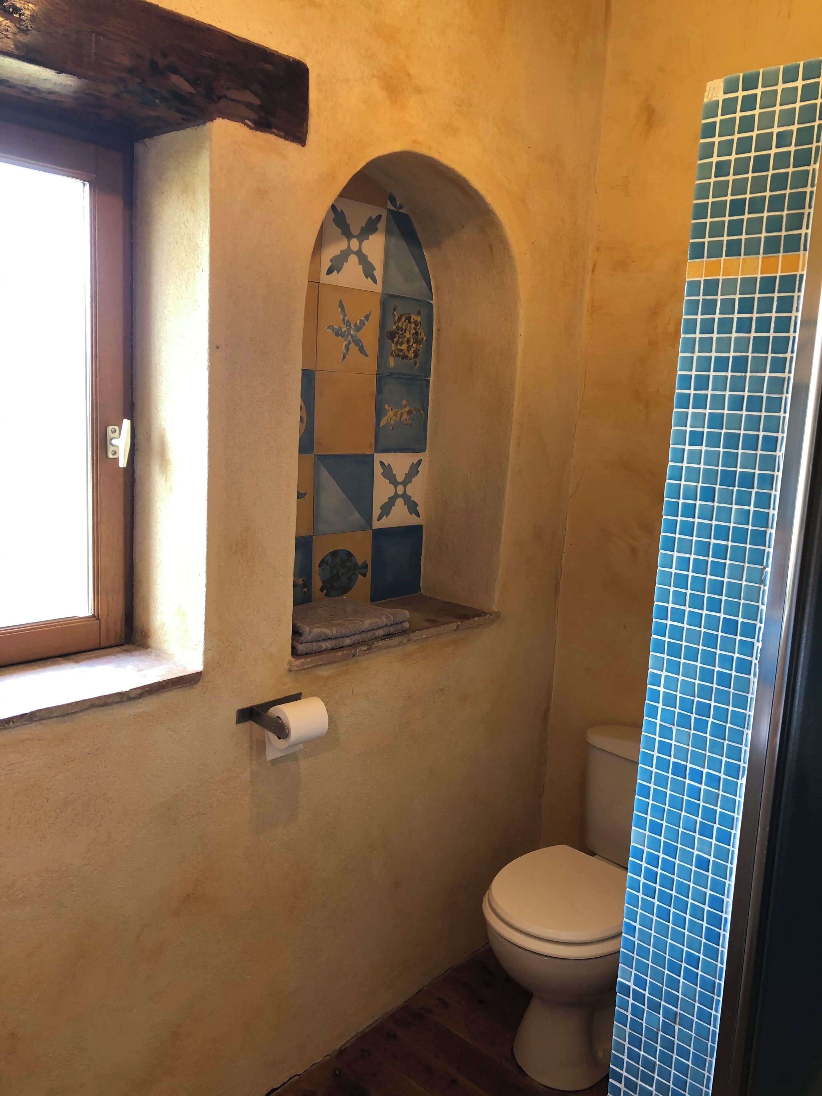 Un baño con pared de azulejos, una ventana, un inodoro y un estante con un rollo de papel higiénico.