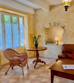 A room with a wicker chair, a wooden table with a vase, a leather sofa, and a stone fireplace. - Gîte Mûrier (Suze-la-Rousse)