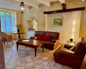 A living room with a brown leather sofa, a wooden coffee table, a wicker chair, and a stone wall with a painting. - Gîte Mûrier (Suze-la-Rousse)