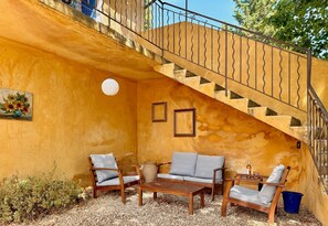 Un espace extérieur aménagé avec des places assises, baigné de lumière naturelle, comprenant une table et des chaises en bois, un luminaire suspendu et un escalier doté d’une rampe en métal.