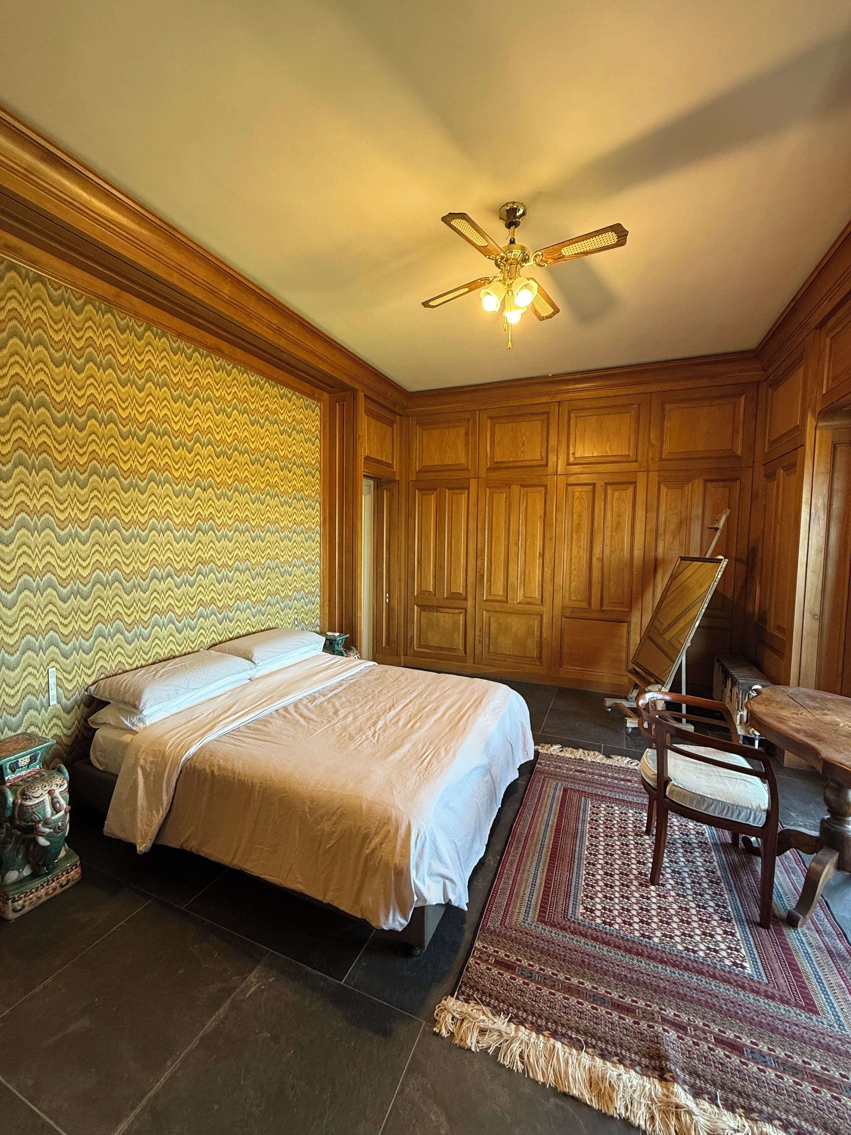 A bedroom with a bed, wooden paneling, a ceiling fan, a chair, and a table.