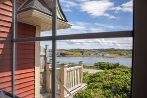 A view through a window of a house with a wooden deck, a small porch, and a view of a body of water with houses in the distance. - Twillingate Little Suite w/ Ocean & Garden View (Twillingate)