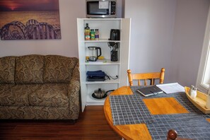 A room with a patterned sofa, a dining table with a placemat, a white shelving unit with kitchen items, and a painting on the wall. - Twillingate Little Suite w/ Ocean & Garden View (Twillingate)