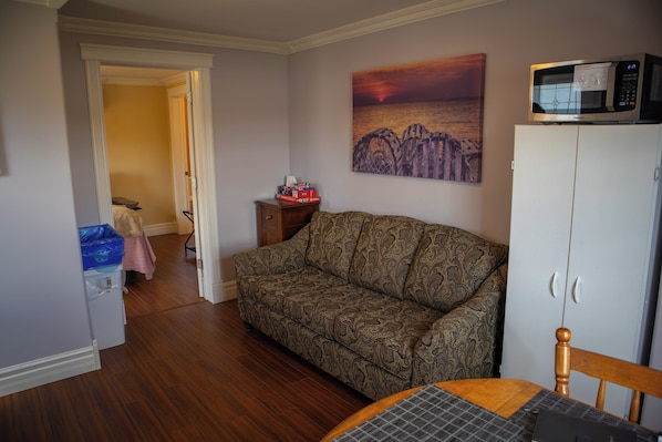 Un salon avec un canapé à motifs, une table à manger recouverte d’une nappe à carreaux, un four à micro-ondes posé sur un meuble et un tableau représentant un coucher de soleil accroché au mur.