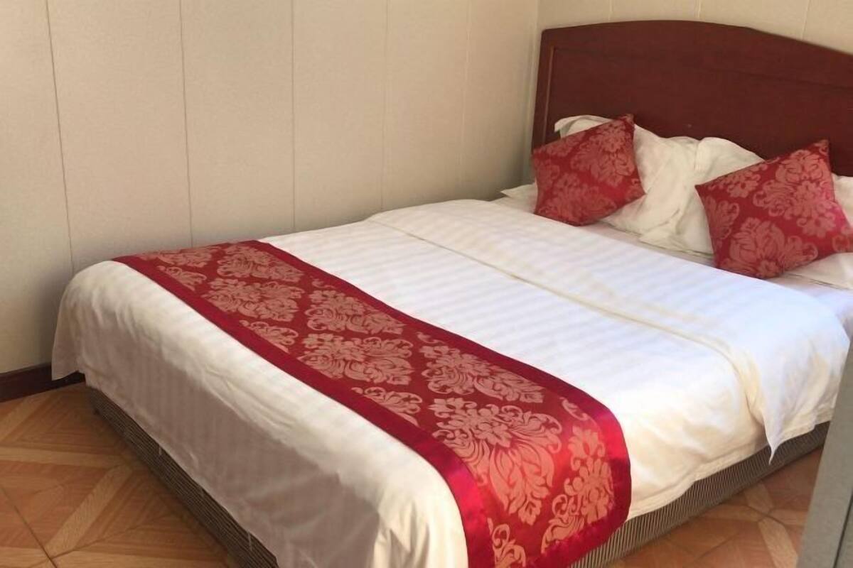 Un lit bien fait, recouvert d’une couvre-lit blanche et rouge assortie aux coussins, se dressant devant un mur sobre et un plancher en bois.