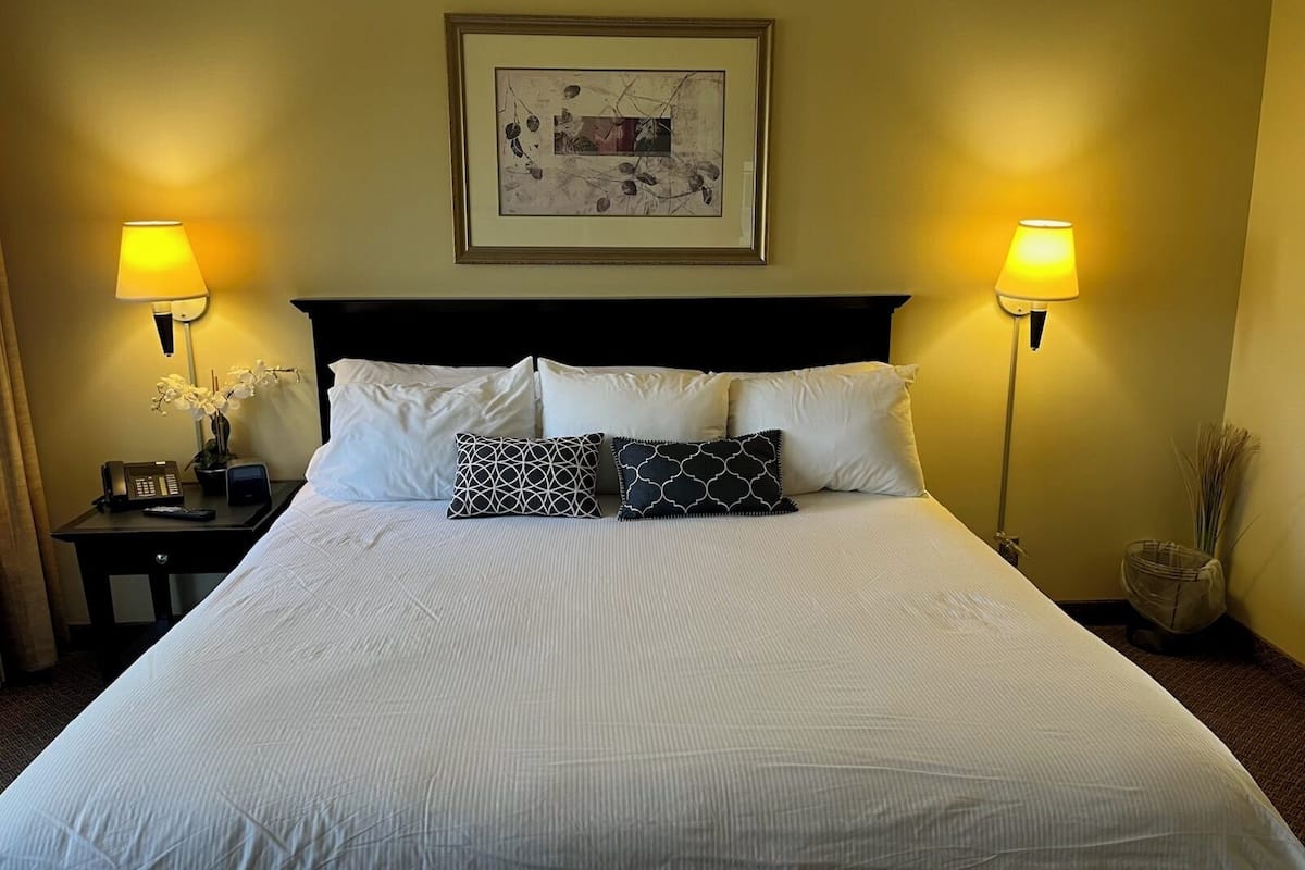 A neatly made bed with white linens and two decorative pillows, flanked by bedside tables with lamps and a telephone.