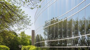 En modern byggnad med en reflekterande glasfasad, omgiven av frodig grönska och en klar himmel.